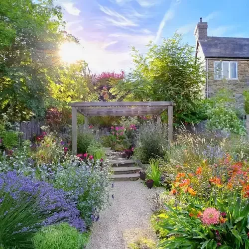 Garden with flowers, gravel path and pergola over steps, with stone house in the background and trees to the left
